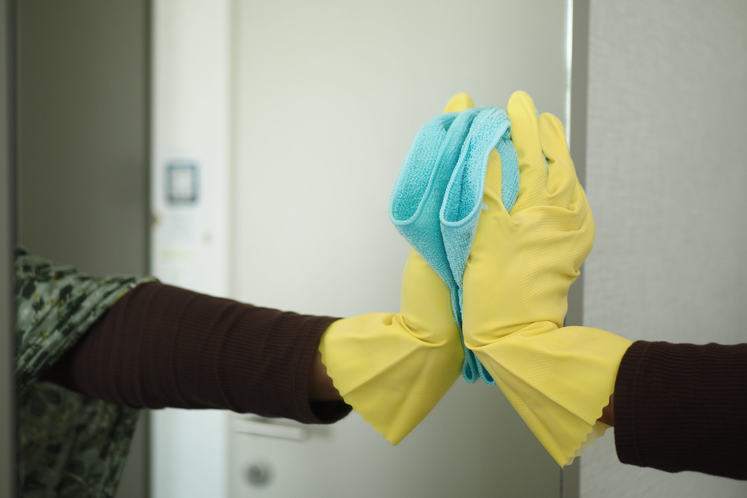 Gloved hands holding a blue microfiber cloth and wiping a white door or cabinet surface in a cleaning scene.